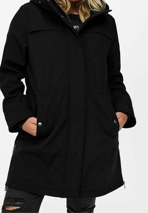 Kvinde iført en lang sort frakke med hænderne i forlommerne og lyse, revnede sorte jeans mod en ensfarvet hvid baggrund.