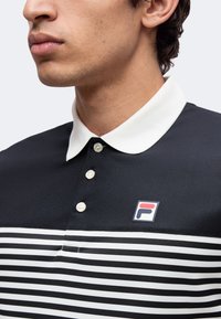 Homme portant un polo rayé noir et blanc avec col blanc et trois boutons, montrant un petit logo rouge et bleu sur la poitrine.