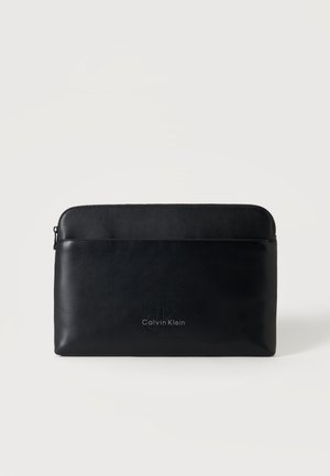 Черен кожен несесер Calvin Klein с цип и ембосирано лого отпред, поставен на светъл едноцветен фон.