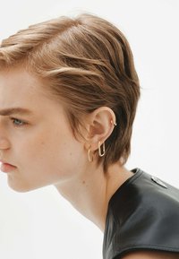 Aros de oro en forma de anillo de diferentes tamaños adornan la oreja. El sujeto tiene el cabello corto y texturizado y lleva una prenda de cuero negra.