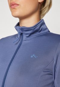 Ljusblå zip-up fleecejacka med hög krage, slät textur och diskret logotypdetalj på bröstet.