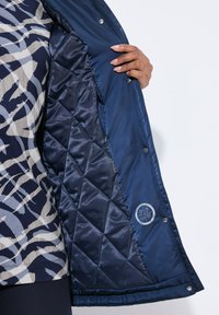 Chaqueta aislada de color navy con un forro interior acolchado. Presenta cierres de presión y un emblema en el interior, combinada con una camiseta de patrón por debajo.