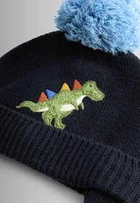 Meresinine kootud müts sinise tutiga, millel on roheline tikitud dinosaurus ja värvilised punased, kollased ning sinised okkad.