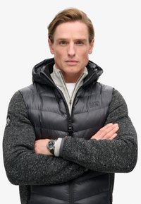 Gilet imbottito nero con un design a maglia grigia, dotato di cappuccio, chiusura con zip e logo sulla spalla sinistra. Accompagnato da una maglia grigia chiara e un orologio.