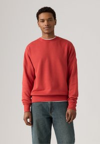 Rode crewneck sweatshirt met ribbelboorden en -zoom, met een subtiele logo op de borst. Gecombineerd met blauwe spijkerbroeken.