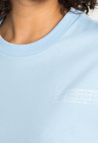 Close-up of a person wearing a light blue T-shirt with white stylized text on the chest reading "Lacoste."