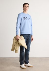 Light blue sweatshirt with black "BOSS" logo, paired with dark denim jeans and white sneakers. Holding a beige jacket with elastic cuffs.