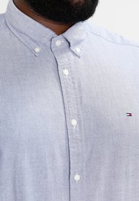 Chemise bleu clair à manches longues en tissu avec une texture subtile. Elle possède un col pointu et un petit logo brodé sur la poitrine.