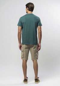 Turkos crewneck-t-shirt, khakifärgade cargoshorts med sidofickor och bruna skor, sedd bakifrån mot en neutral bakgrund.