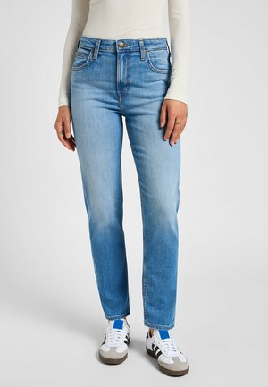 Žena nosící světle modré džíny s rovnými nohavicemi, offwhite žebrovaný top s dlouhými rukávy a bílé tenisky s černými pruhy a modrými akcenty.