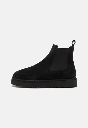 Botas de tornozelo em camurça preta com painéis laterais elásticos e um tab do tipo laço. Apresenta uma sola de borracha preta grossa com uma borda serrilhada para maior tração.