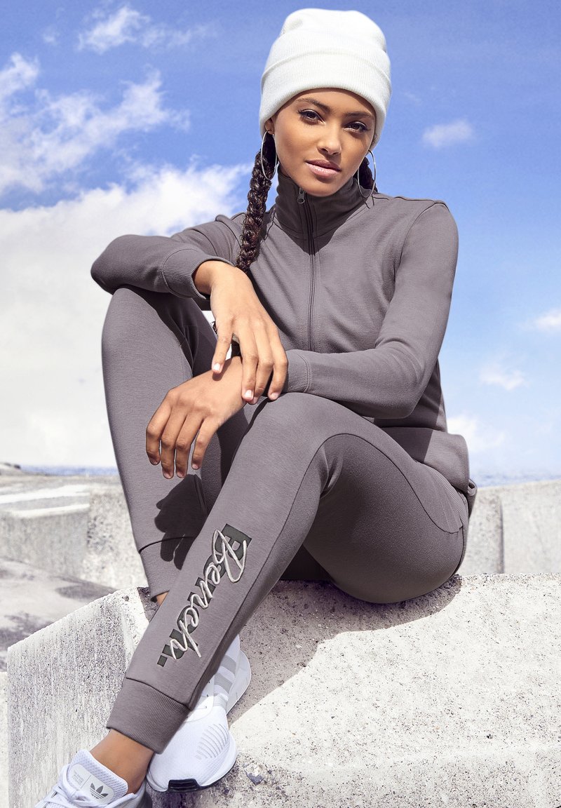Grauer Sportanzug mit einem Reißverschluss-Oberteil, dazu passende Leggings mit Logodetail und einer weißen Mütze, die auf einer Betonoberfläche sitzt.