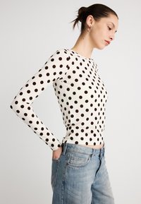 Pitkähihainen valkoinen paita mustilla pilkuilla, istuva malli, pyöreä pääntie, yhdistetty vaaleansinisiin farkkuihin, joissa on viiden taskun rakenne.