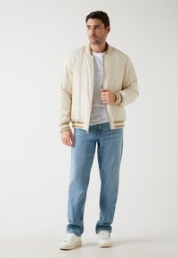 Beige prošívaná bomber bunda s pruhovanými manžetami, nošená přes bílé tričko a světle modré džíny, spárovaná s bílými teniskami.