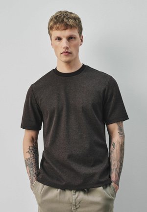 Jeune homme aux cheveux courts portant un T-shirt texturé foncé et un pantalon beige, debout les mains dans les poches sur un fond clair uni.