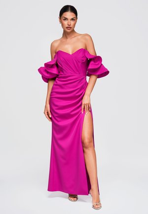 Femme portant une robe rose vif épaules dénudées avec des manches à volants et une haute fente, debout devant un fond clair uni.