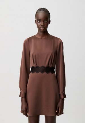 Mujer con el cabello trenzado vistiendo un vestido marrón de manga larga con una cintura de encaje negro, de pie frente a un fondo gris claro y liso.