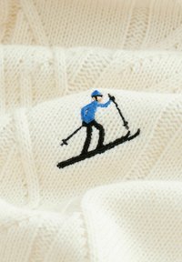 Knit krēmkrāsas džemperis ar zilu slēpotāju izšuvumu uz priekšpuses. Auduma struktūra ir mīksta ar ribotu rakstu.