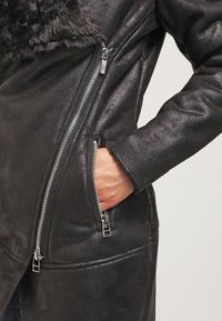 Abrigo de cuero negro con un acabado texturizado, que presenta un cuello de piel y cremalleras plateadas. Los bolsillos tienen cremalleras dobles para un detalle adicional.