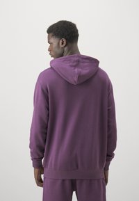 Sudadera morada con capucha, de mangas largas y puños acanalados, que presenta un corte relajado y una textura suave. Costuras detalladas visibles en las uniones.