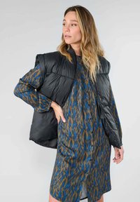 Gilet matelassé noir sur une robe bleue et marron à motifs, avec un devant à boutons, des manches longues et une légère fente à l'ourlet.