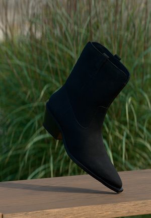 Schwarzer Wildleder-Stiefelette mit niedrigem Absatz, der am Rand einer Holzoberfläche ruht, mit grünem, hohem Gras im Hintergrund.
