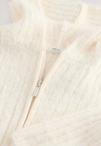 Pull à fermeture éclair couleur crème en tissu tricoté doux et texturé. Doté d'une capuche et d'un subtil motif côtelé vertical. Étiquette visible.