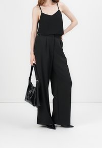 Camiseta negra sin mangas con tirantes finos, combinada con pantalones anchos negros de cintura alta. En una mano se sostiene un bolso de cuero negro con correa.
