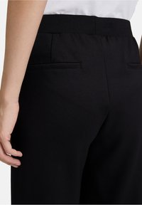 Pantalon noir avec une texture de tissu lisse, une taille élastique et deux poches arrière avec une finition soignée, présenté de côté.