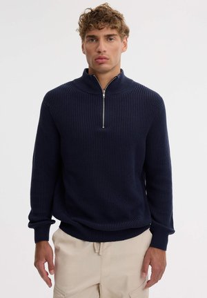 Un hombre posa con un jersey azul marino de punto acanalado y cremallera hasta el pecho, combinado con unos pantalones color crema, sobre un fondo liso y claro.