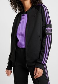 Svart zip-up jacka med lila ränder på ärmarna och en asymmetrisk skärning, kompletterad med en matchande lila ribbad topp under.