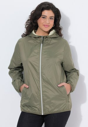 Vrouw met krullend donker haar, gekleed in een olijfgroene capuchontrui met rits en handen in de zakken, glimlachend tegen een effen lichte achtergrond.