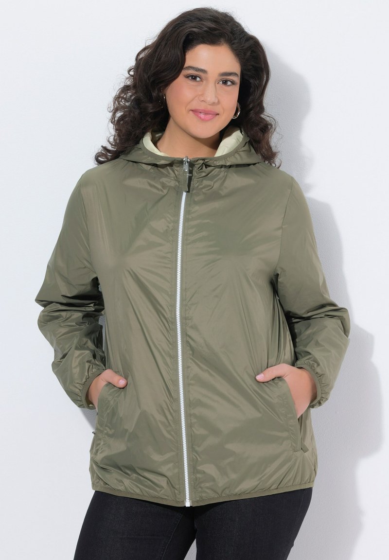 Kobieta z kręconymi ciemnymi włosami, ubrana w oliwkowo-zieloną kurtkę z suwakiem i kapturem, z rękami w kieszeniach, uśmiechająca się na jasnym tle.