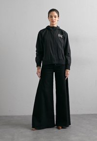 Casaco corta-vento preto com fecho frontal, punhos elásticos e logótipo bordado. Combinado com calças largas pretas, ambos feitos de tecido leve.