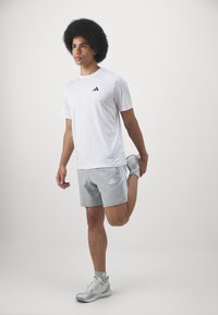 T-shirt de sport blanche avec logo noir, short gris avec rayures blanches, chaussures de course grises. Sujet s'étirant une jambe vers l'arrière.