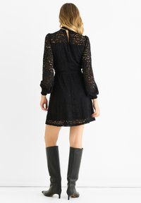 Black lace dress with long sleeves, fitted waist, and flared skirt; features a keyhole back and textured floral pattern. Paired with knee-high black boots.