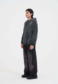 Sudadera con cremallera de color gris oscuro con tachuelas, combinada con pantalones negros holgados que presentan intrincados patrones de serpiente. El modelo se encuentra de pie frente a un fondo blanco.