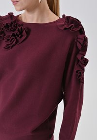 Maglione a maglia bordeaux con dettagli arricciati sulle spalle e scollatura rotonda. Tessuto morbido, vestibilità comoda e maniche lunghe.