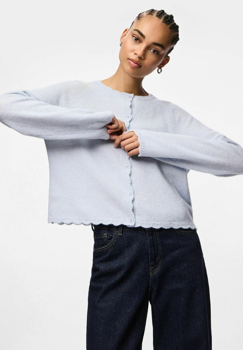 Light blue cropped cardigan made from soft material, featuring a scalloped hem, round neckline, and button closure. Worn with dark jeans.