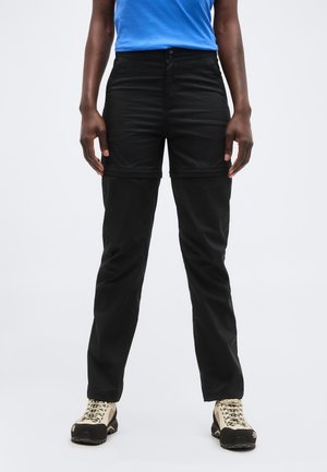 Άτομο που φοράει μαύρο παντελόνι πεζοπορίας με φερμουάρ και μπεζ-μαύρα παπούτσια πεζοπορίας, στέκεται μπροστά σε απλό λευκό φόντο.