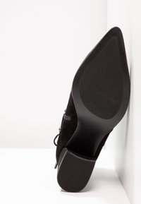 Scarpa nera in suede con tacco alto e punta affilata, suola in gomma testurizzata e lacci decorativi ai lati. Dettagli di cucitura chiari visibili.
