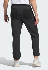 Vyras, vilkintis juodus sportinius kelnes su užpakalinėmis kišenėmis ir baltus sportinius batelius, stovi prieš paprastą šviesų foną.