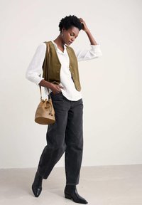 Maslinasto zeleni kordurojski prsluk preko bijele bluze, crne široke traperice i crne gležnjače. Smeša kožna torba s uzicom dodaje detalj.