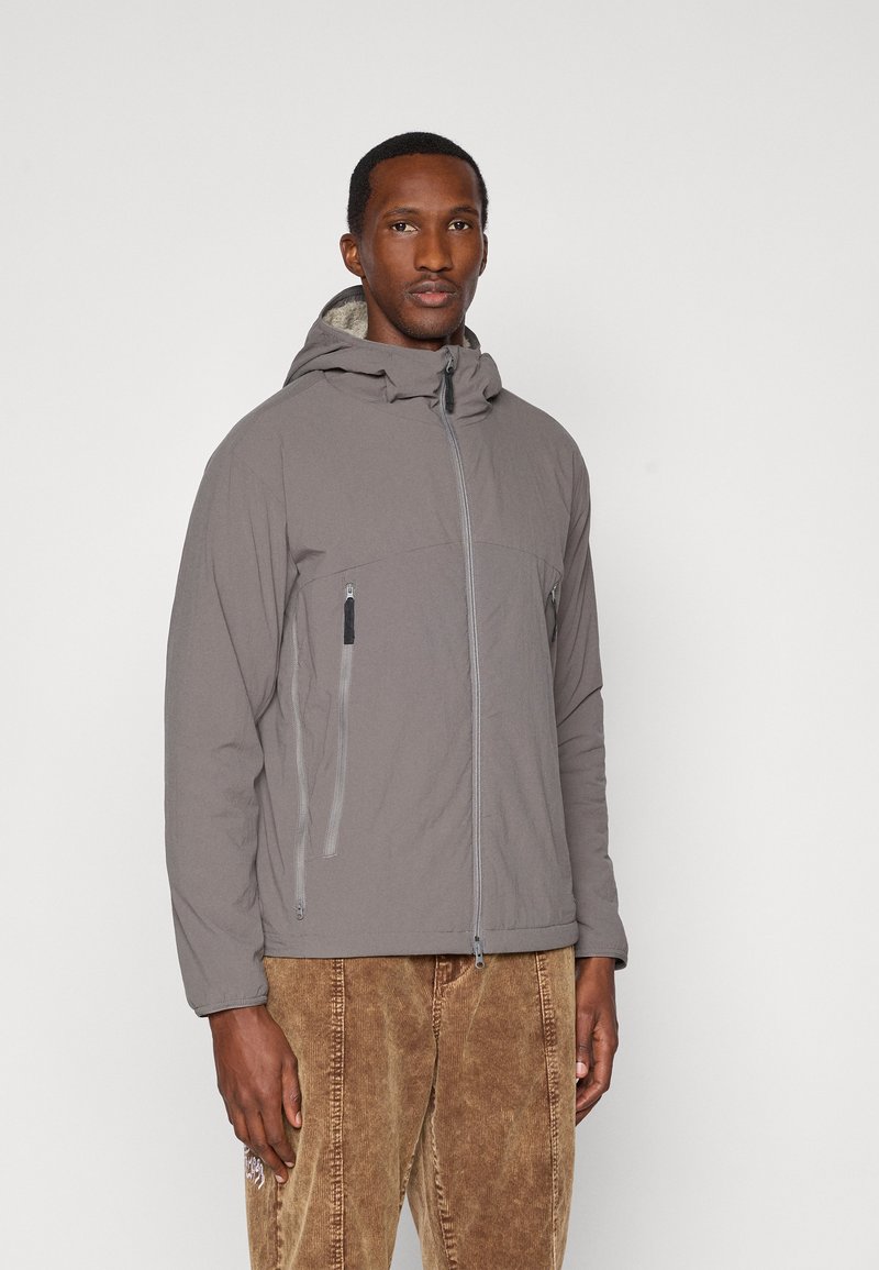 Veste grise claire avec une fermeture éclair à l'avant, une capuche et deux poches latérales, présentant une texture lisse et un design minimaliste, associée à un pantalon en velours côtelé marron.