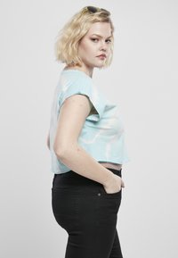 Svetlomodré skrátené tričko s bielym tie-dye vzorom, krátke rukávy, skombinované s čiernymi džínsami, model stojí s rukou v vrecku.