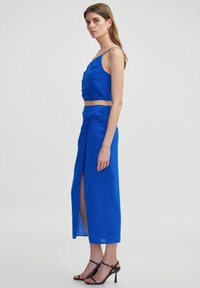 Twee-delig outfit in felblauw. Het topje heeft een geknoopt ontwerp met dunne bandjes, terwijl de rok lang is, gerimpeld bij de taille, met een split aan de zijkant.