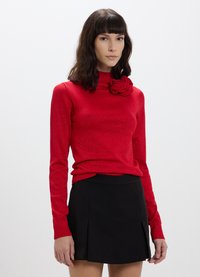 Maglione rosso a maniche lunghe con colletto alto e un dettaglio in rosa testurizzato sul petto, abbinato a una gonna nera plissettata.