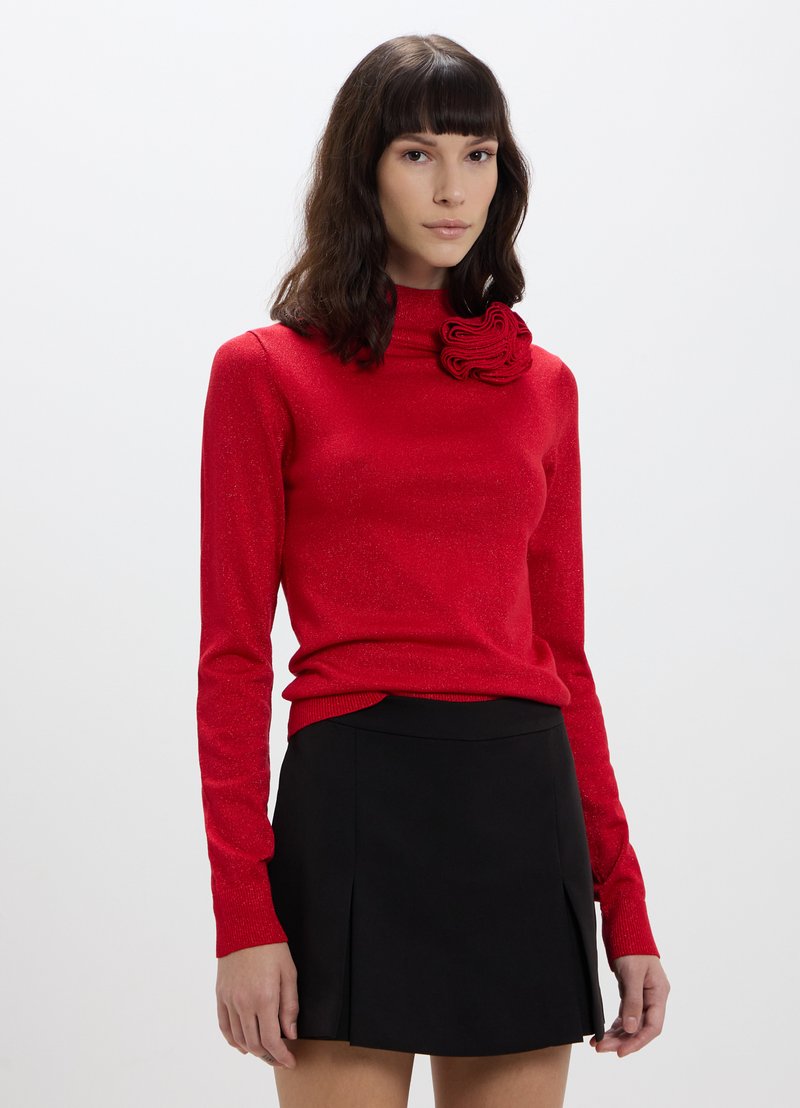 Maglione rosso a maniche lunghe con colletto alto e un dettaglio in rosa testurizzato sul petto, abbinato a una gonna nera plissettata.