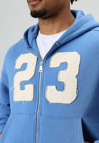 Blauwe hoodie met rits en grote beige versleten stoffen cijfers "23" aan de voorkant. Voorzien van een kangoeroezak en een capuchon met trekkoord.