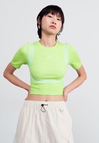 Trumpas rankovėmis laisvas trumposios palaidinės modelis ryškiai kalkių žaliai. Lygus audinys su tinklinėmis detalėmis ant pečių. Įrengtas balta Adidas logo krūtinėje.
